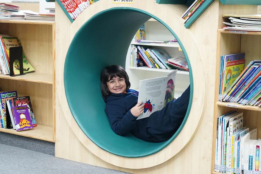 A primary school student is reading a book in a cozy spot at the Prometheus School library.