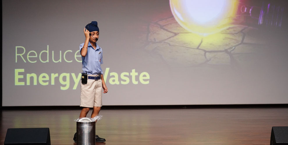 A student from Prometheus School is giving presentation on stage. The background displays the text "Reduce Energy Waste," highlighting the school's commitment to teaching students about energy conservation and sustainability practices