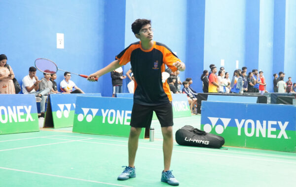 Serving-A Prometheus School student prepares to serve, tossing the shuttlecock into the air with a focused stance on the badminton court