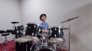 Student in a school uniform playing a black drum set in a music room, with an electronic drum kit in the background.