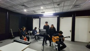 Students performing in a music rehearsal, with a singer, guitarist, keyboardist, and percussionist in a soundproof room.