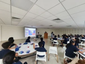 Students seated at round tables attending an interactive session with a large screen presentation at the front, while a speaker addresses the group from a podium.