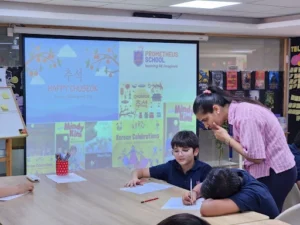 Teacher assisting young students during a classroom activity about Chuseok, Korean Thanksgiving Day, with a presentation screen displaying festive visuals and books in the background