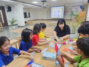 Teacher guiding a group of young students in an origami activity at a classroom table, with a Chuseok-themed presentation displayed in the background.